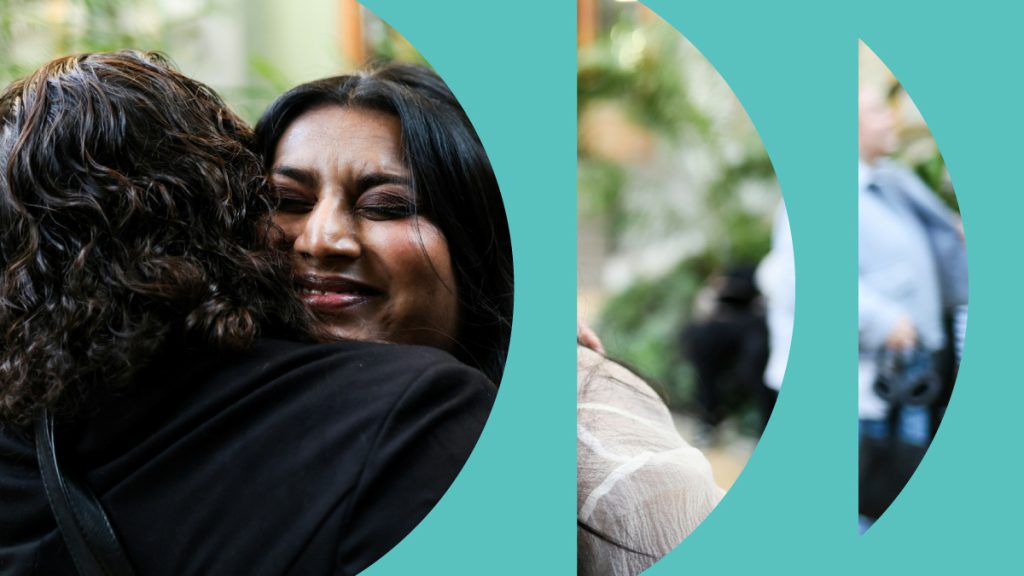 Two women embracing in a warm hug, eyes closed, split across teal circular design elements. Part of the Intention 2 Impact "Impact is a Verb" campaign.